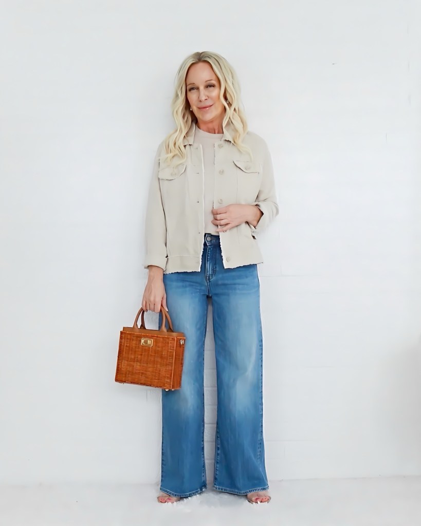 A woman with long blonde hair is standing against a white background, wearing a light beige jacket and wide-leg blue jeans. She holds a small wicker handbag and is smiling slightly.