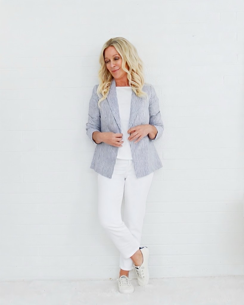 A woman with long, wavy blonde hair wearing a striped blazer over a white shirt, paired with white pants and sneakers, standing against a white wall.