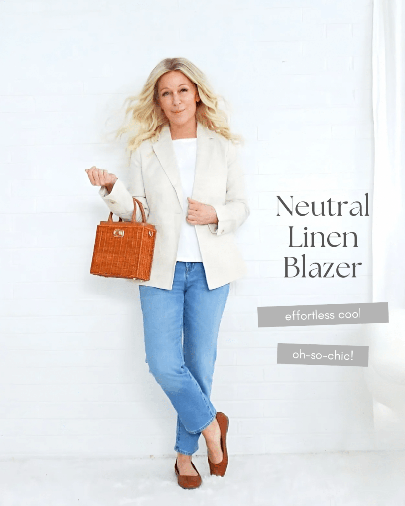 A woman wearing a neutral linen blazer, white top, and blue jeans, posing with a woven handbag in front of a white wall.