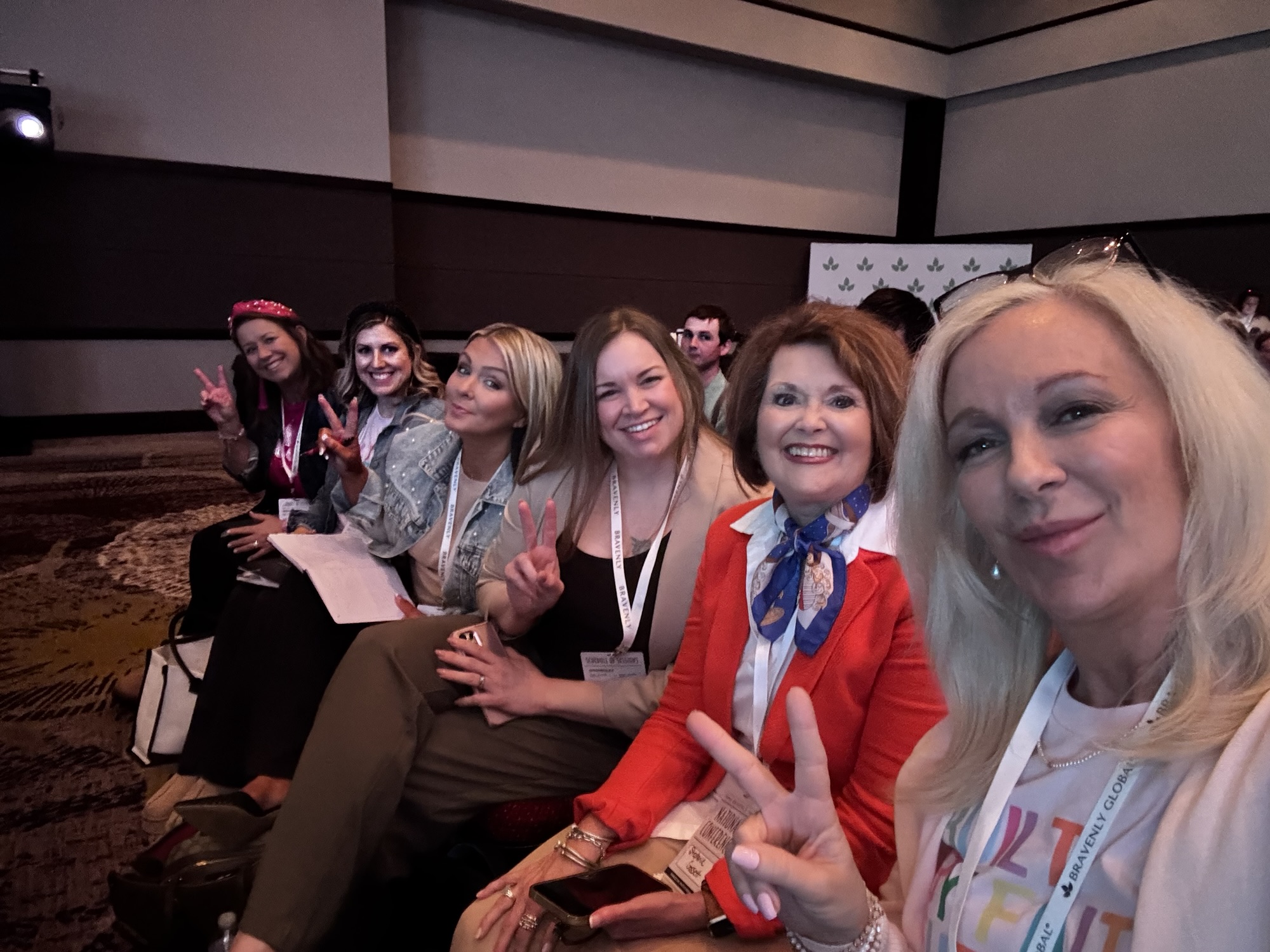 A group of smiling women sitting together, posing with peace signs at a conference.