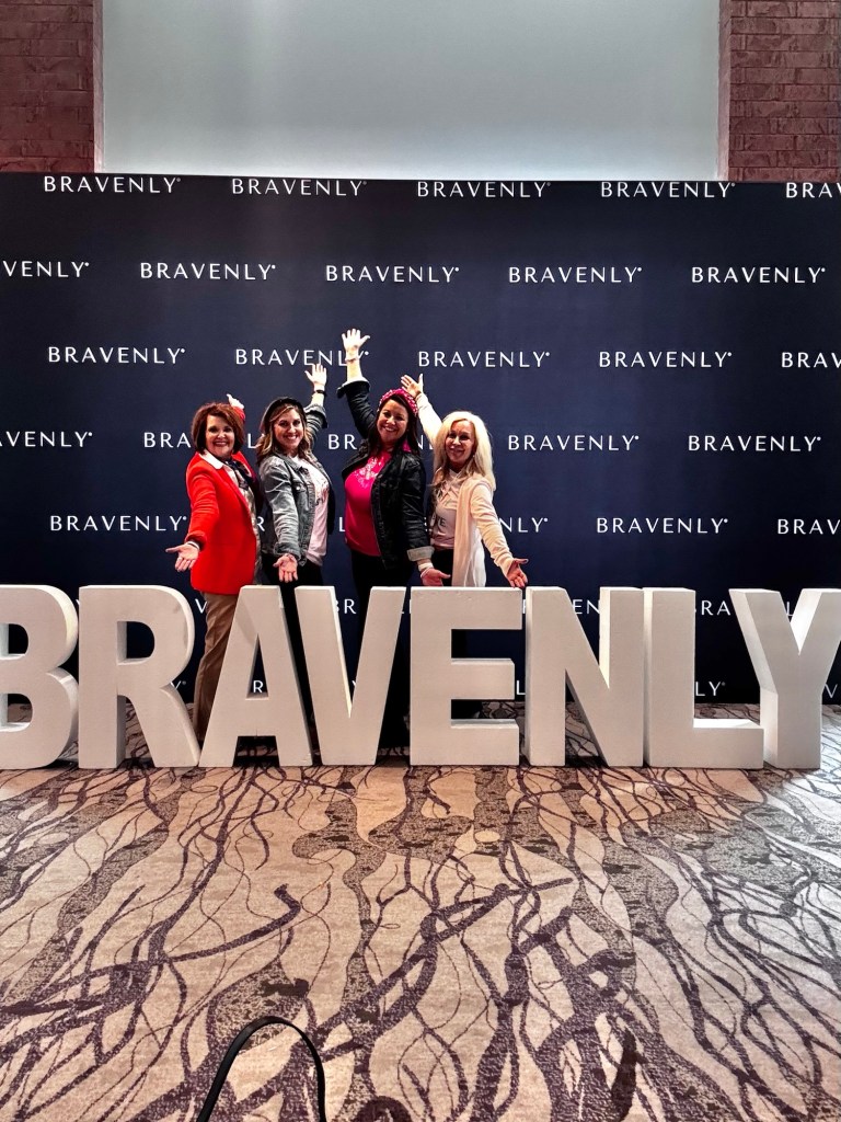 Four women posing happily in front of large letters spelling 'BRAVENLY' at an event, with a branded backdrop.