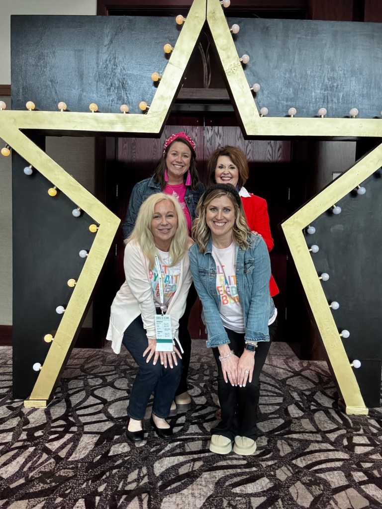 Four women posing together in front of a large star decoration, with lights attached. They are smiling and dressed casually, showcasing a lively atmosphere.