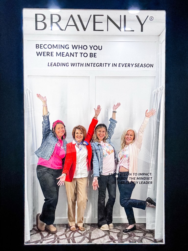 Four women joyfully posing together with raised arms, wearing colorful clothing and smiling, in front of a backdrop with the text 'BRAVENLY' and inspirational messages.
