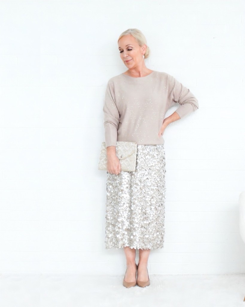 A woman wearing a light beige sweater and a shiny sequined skirt, holding a matching clutch bag, posed against a white background.