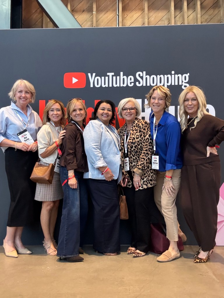 A group of seven women standing together in front of a large YouTube Shopping banner at an event.