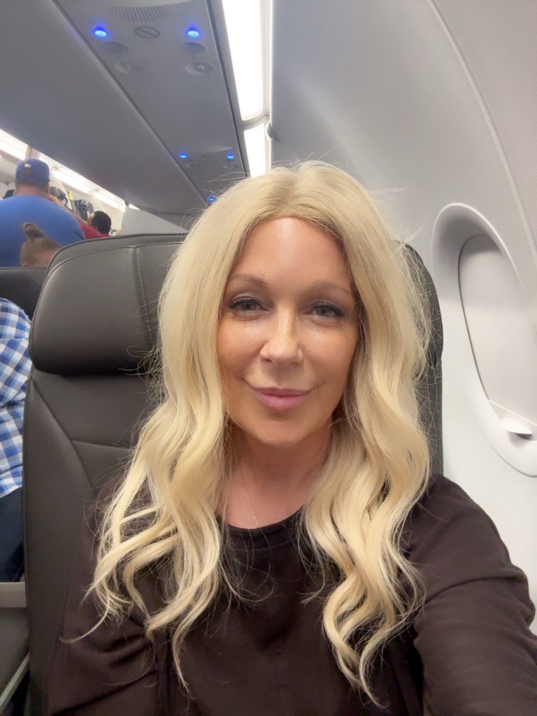 A person with long blonde hair sitting in an airplane seat, smiling at the camera. The interior of the aircraft is visible, featuring overhead lights and other passengers in the background.