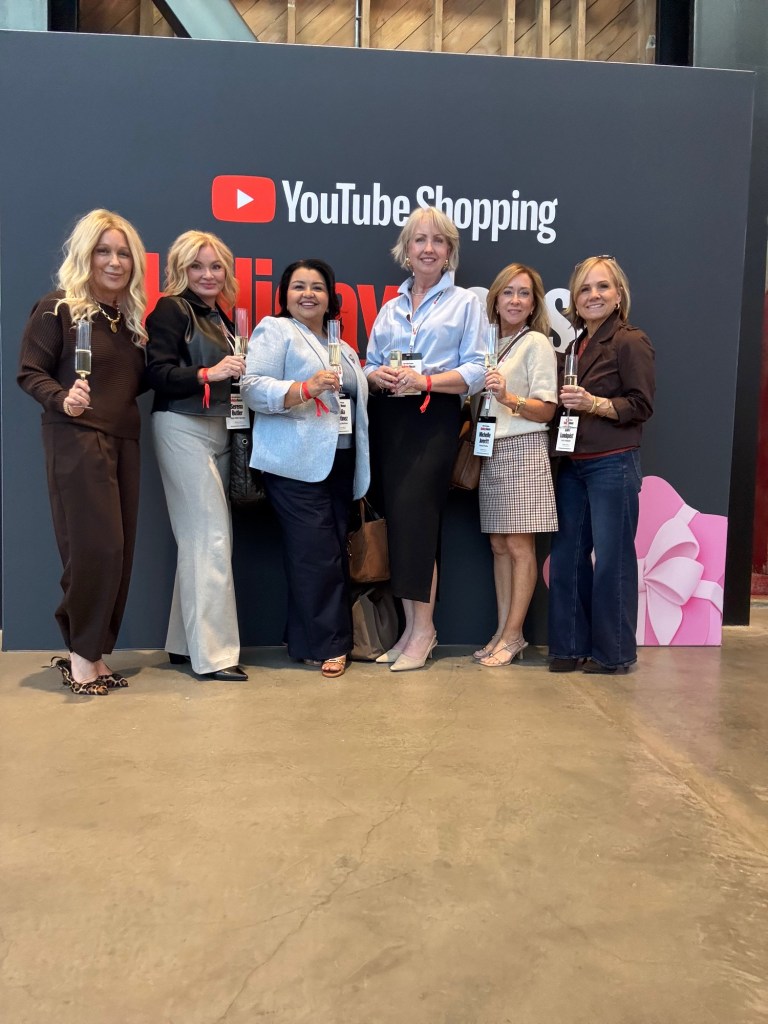 Group of six women posing together at a promotional event for YouTube Shopping, holding drinks and standing in front of a large YouTube Shopping backdrop.