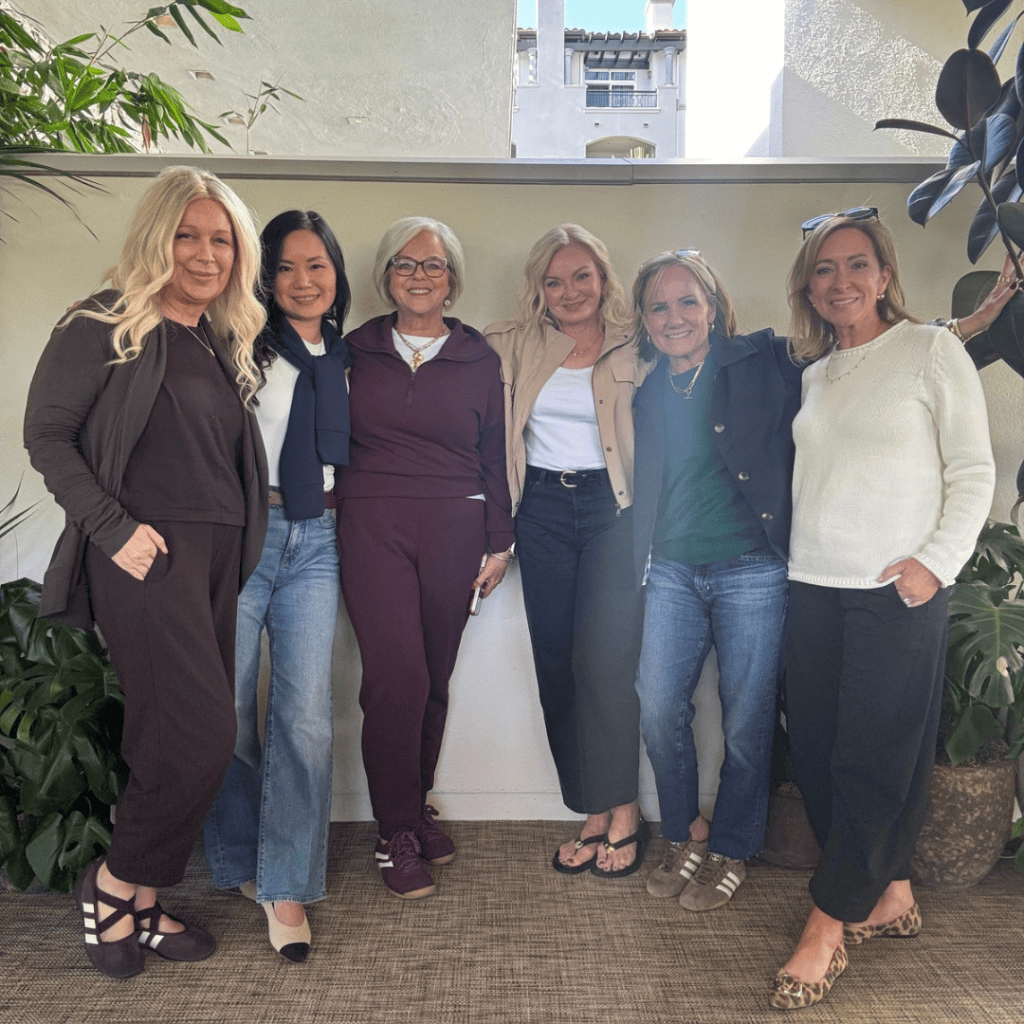 A group of six women posing together in a casual and friendly setting, standing in front of greenery and a wall.